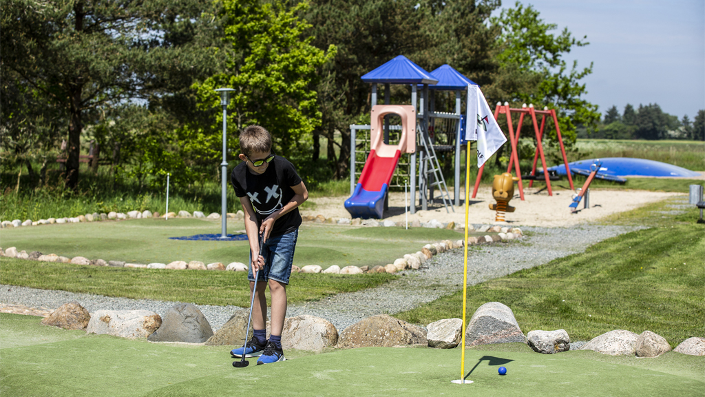 Dreng putter med legepladsen i baggrunden på Lindvig Minigolf